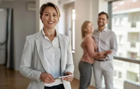 Imagem de uma corretora sorridente, especializada em administração de imóveis, está em primeiro plano segurando um tablet, enquanto um casal feliz se abraça ao fundo, próximo a uma grande janela em um apartamento moderno e bem iluminado.