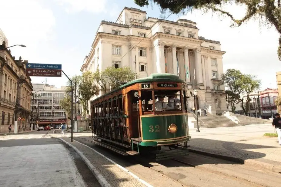 Imagem de um bonde verde, com o número 32, percorre os trilhos em frente a um grande edifício histórico com colunas em Santos