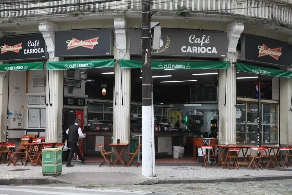 Imagem da vista externa do Café Carioca, em Santos, com mesas e cadeiras na calçada e um garçom atendendo os clientes. Toldos verdes e colunas emolduram a fachada do café.