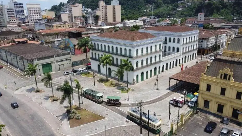 Imagem da vista aérea de um prédio histórico branco com telhado vermelho, palmeiras e ruas de paralelepípedos no Centro Histórico de Santos. Um bonde verde e branco passa nas proximidades, cercado por outros prédios da cidade e por um pouco de vegetação ao fundo.