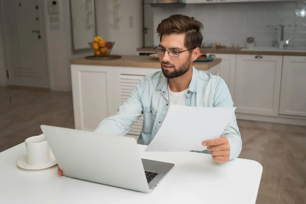 Imagem de um homem de óculos sentado em uma mesa de cozinha, olhando para um laptop e segurando uma folha de papel, para ilustrar matéria sobre crédito imobiliário com garantia de imóvel. Uma xícara e um pires estão sobre a mesa; uma fruta pode ser vista no balcão ao fundo.