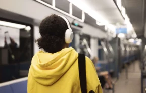 Imagem de uma pessoa usando um capuz amarelo e fones de ouvido brancos em um trem de metrô moderno, quase vazio, com assentos azuis, para ilustar matéria sobre a Linha Amarela, do metrô de SP