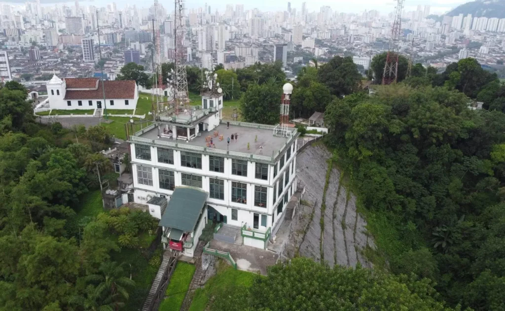 Imagem da vista aérea do Monte Serrat, em Santo, com um prédio branco situado na encosta, com antenas, cercado por árvores e com vista para a paisagem urbana. Um funicular leva até o prédio, e uma igreja é visível nas proximidades.