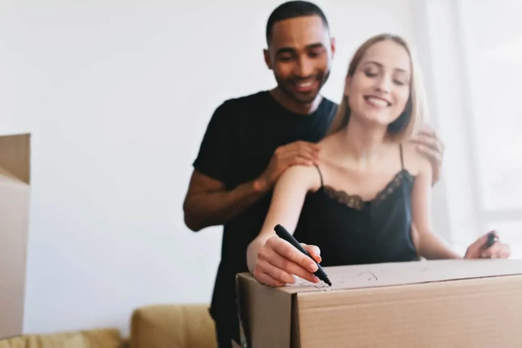 Imagem de um casal sorridente se preparando para uma mudança, com a mulher escrevendo em uma caixa de papelão usando um marcador, enquanto o homem fica atrás dela com as mãos em seus ombros.