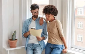 Imagem de um casal formado por um homem e uma mulher em pé encostados na janela de uma casa olhando para documentos que estão na mão do homem para ilustrar matéria sobre o que é usucapião