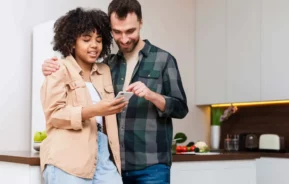 Imagem de um casal sorridente em uma cozinha moderna, olhando juntos para um smartphone, para ilustrar matéria sobre permuta de imóveis. A mulher segura o telefone enquanto o homem a abraça, com legumes frescos visíveis no balcão atrás deles.