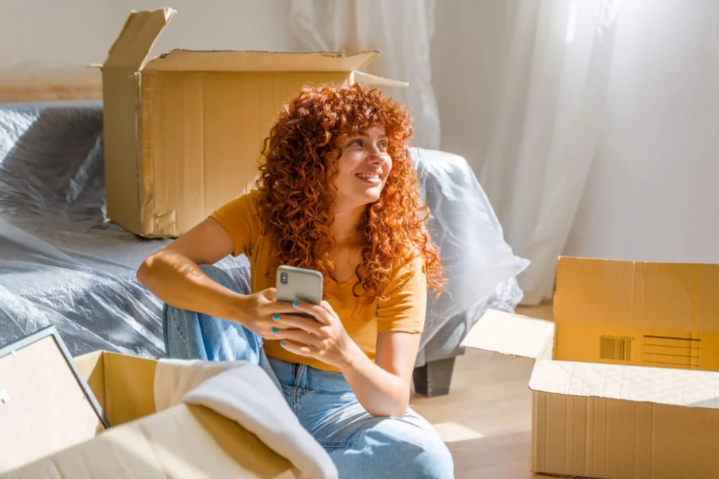 Imagem de uma mulher de cabelos ruivos encaracolados sentada no chão, cercada por caixas de mudança abertas, segurando um smartphone e sorrindo para ilustrar matéria sobre as regras para saque do FGTS para compra de imóvel. A luz do sol entra por uma janela e um sofá coberto está ao fundo