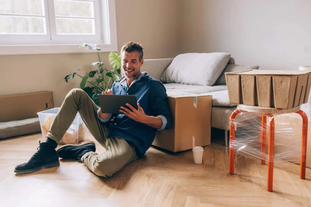 Imagem de um homem sentado no chão de uma sala de estar entre caixas de mudança, sorrindo enquanto usa um tablet, para ilustrar matéria sobre as regras para uso do FGTS na compra de imóvel. Há um sofá, uma planta e uma xícara de café por perto; alguns móveis ainda estão embrulhados em plástico