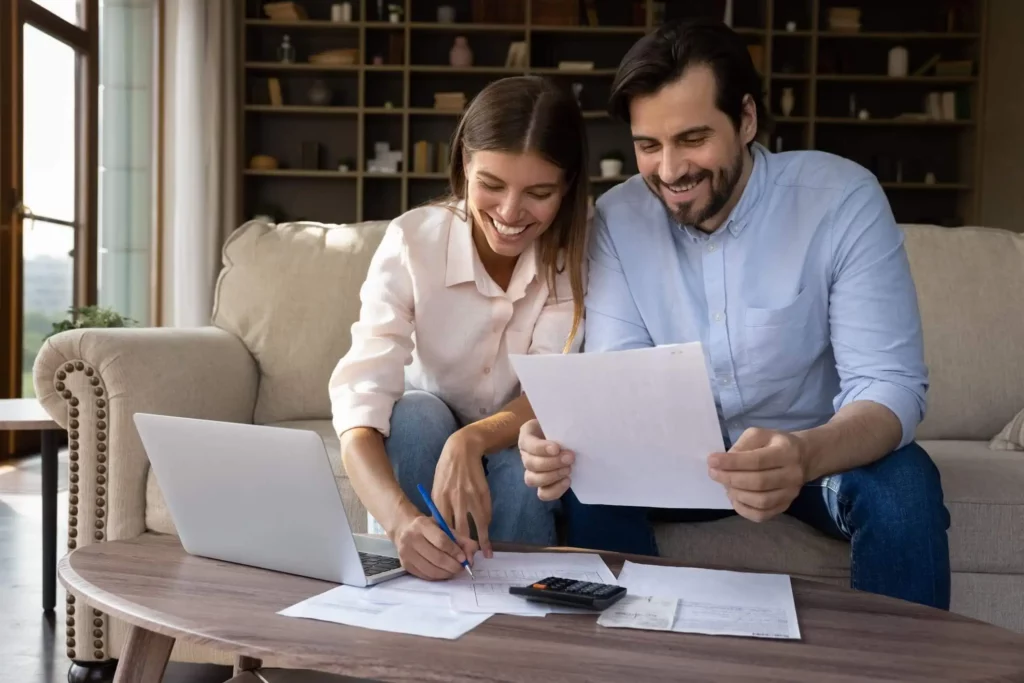Imagem de um casal sorridente sentado em um sofá, trabalhando juntos nas finanças para ilustrar matéria sobre as regras para utilização do FGTS para compra de imóvel. Eles têm papéis, uma calculadora e um laptop à sua frente, parecendo concentrados e felizes. Ao fundo, há prateleiras com livros