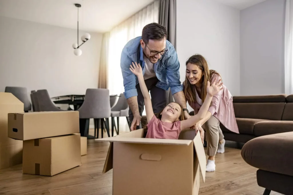 Imagem de um casal sorridente brincando com sua filha pequena, que se senta alegremente em uma caixa de papelão com os braços levantados cercada por caixas de mudança em uma sala de estar moderna e iluminada para ilustrar matéria sobre o uso do FGTS para compra de segundo imóvel