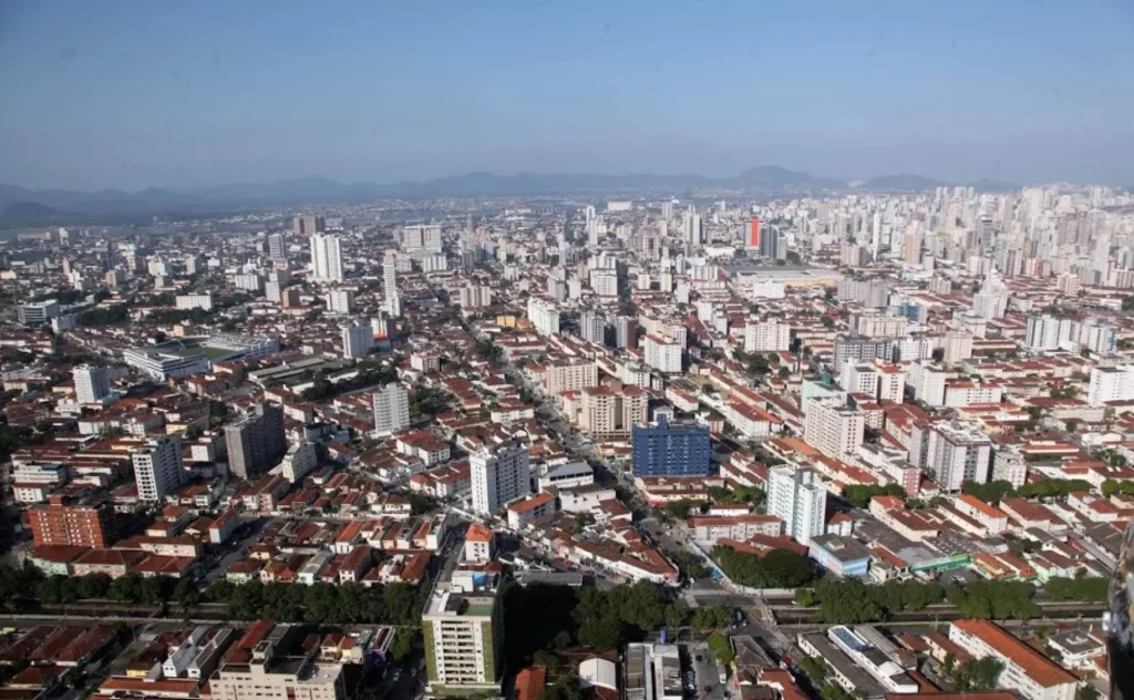 Imagem de vista aérea de Santos, mostra altos e baixos, dispostos de forma compacta, sob um céu azul límpido e com o mar ao fundo.
