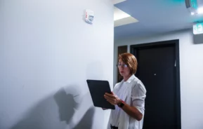 Uma pessoa usando óculos e uma camisa branca está em um corredor, segurando um tablet durante uma vistoria de entrada, olhando para um detector de fumaça montado na parede.