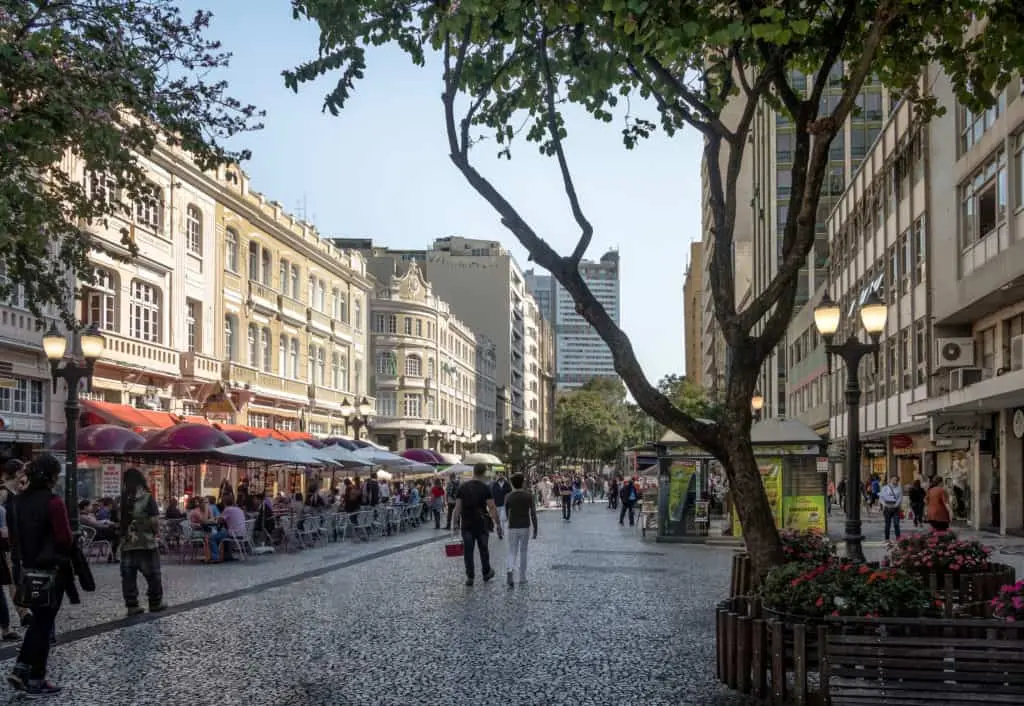 Uma rua de pedestres em um dos melhores bairros de Curitiba para universitários, repleta de prédios históricos, cafés ao ar livre com guarda-sóis vermelhos, pessoas passeando e árvores frondosas que fornecem sombra em um dia ensolarado.