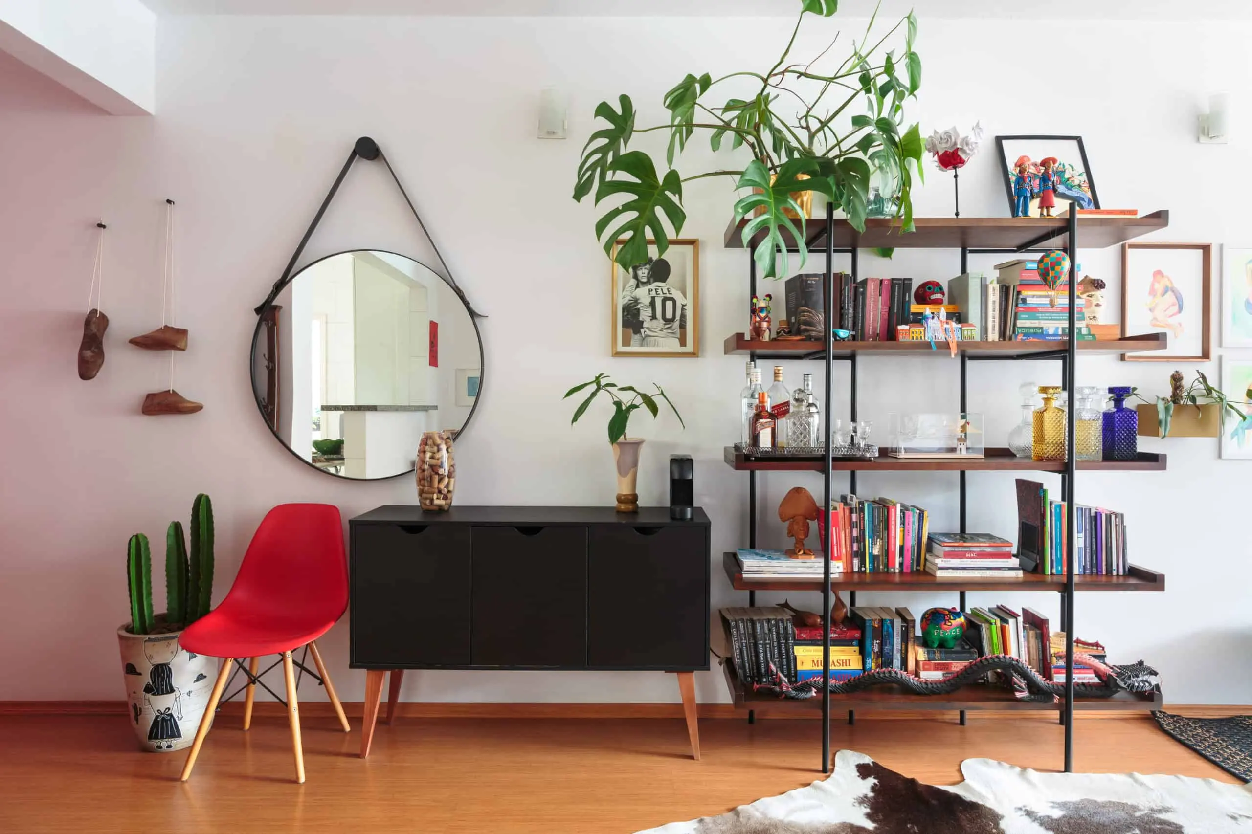 Uma sala de estar moderna no apartamento no Butantã apresenta um espelho redondo sobre um aparador preto, uma cadeira vermelha, vasos de plantas, um cacto e uma estante aberta repleta de livros, decoração e obras de arte emolduradas sobre um piso de madeira clara.