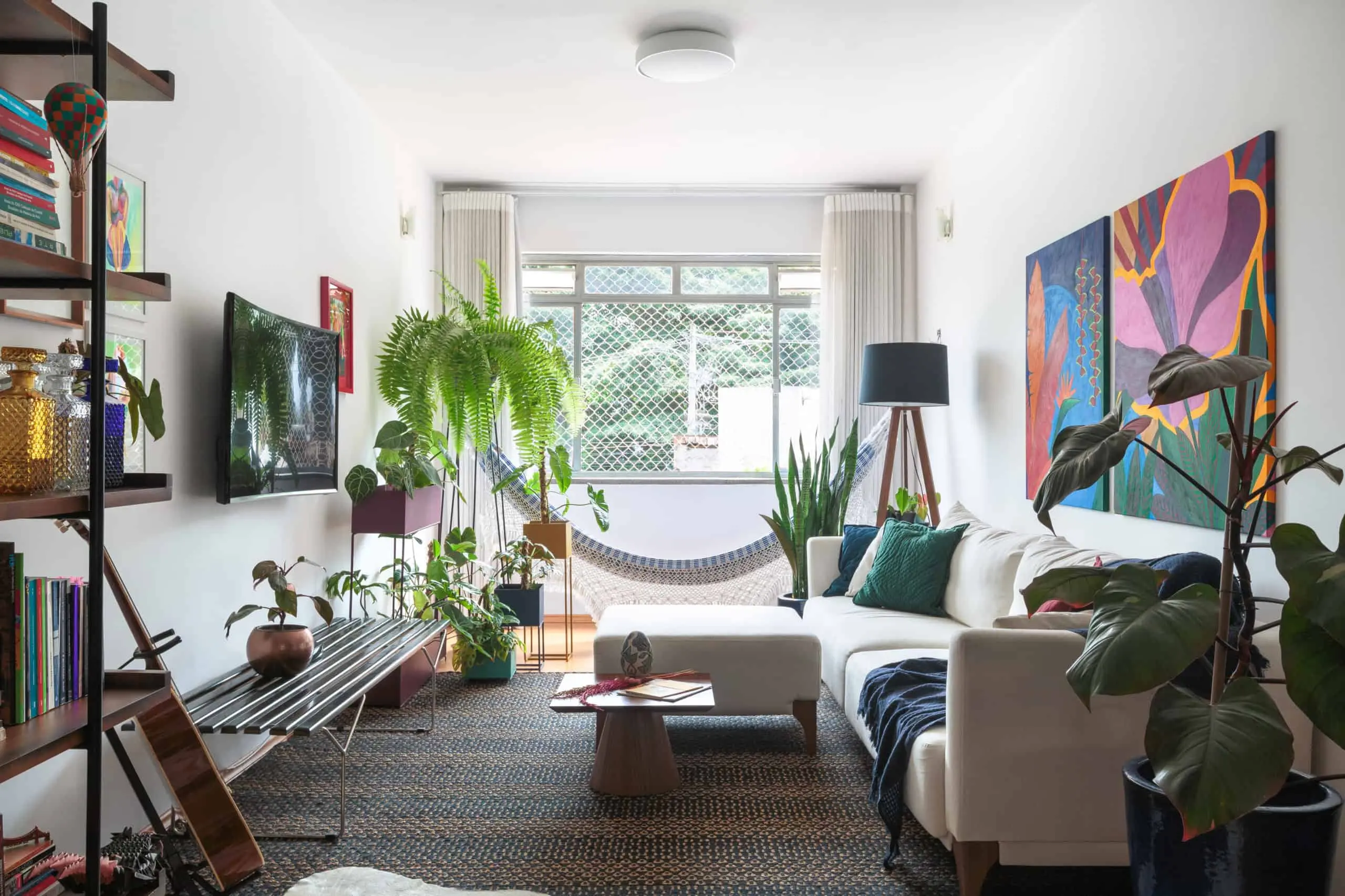 Sala de estar iluminada no apartamento no Butantã com um sofá secional branco, plantas de interior, arte abstrata colorida nas paredes, estantes de livros, uma TV montada na parede e uma grande janela que deixa entrar luz natural.