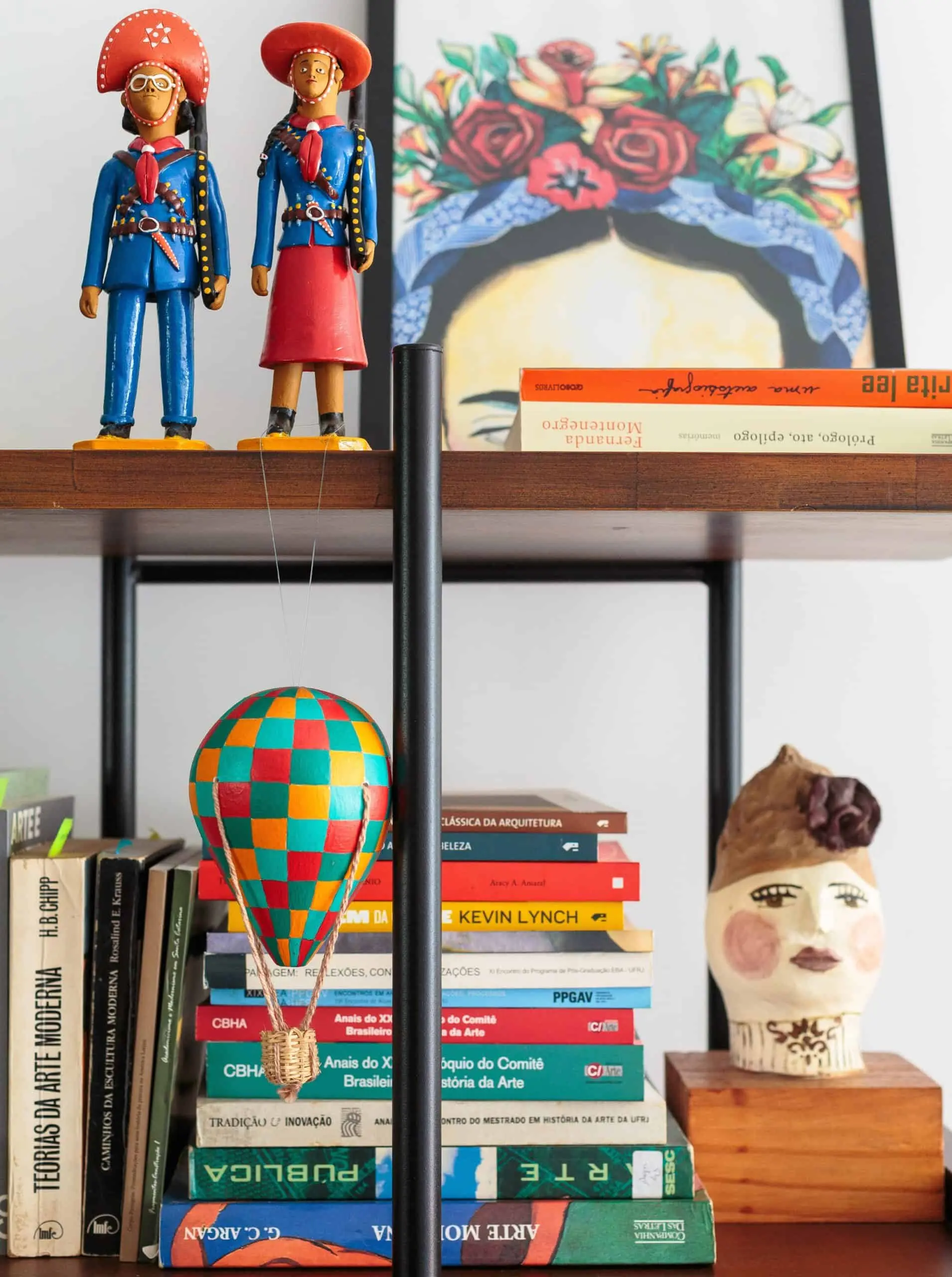 Uma estante em um apartamento no Butantã exibe estatuetas coloridas, um retrato emoldurado de Frida Kahlo, um modelo de balão de ar quente, livros empilhados e uma escultura de cabeça de cerâmica feita à mão, criando um arranjo vibrante e artístico.