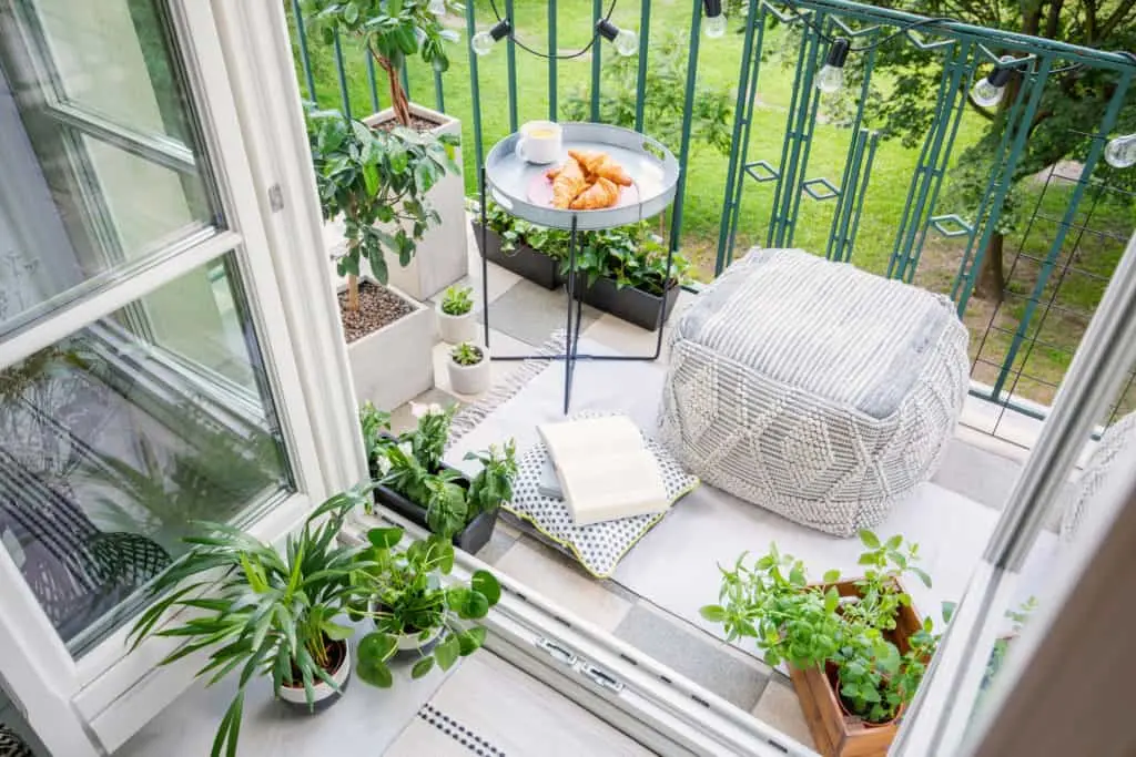 Uma aconchegante varanda com espaço lounge, vasos de plantas, um pufe com um livro e um cobertor, e uma mesa redonda com croissants e uma bebida. Uma grade verde oferece vista para a área gramada externa.