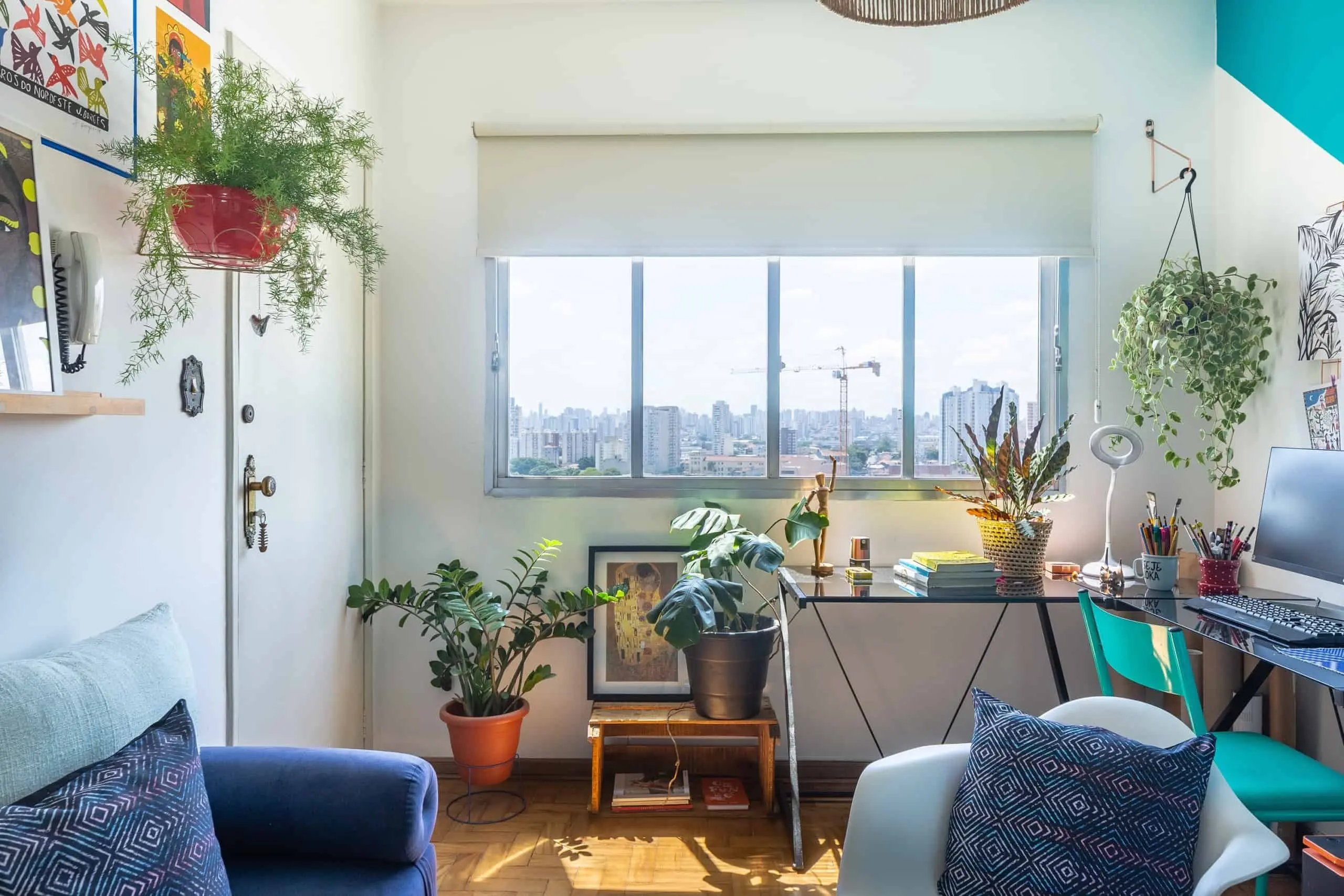 Sala de estar iluminada com diversos vasos de plantas, uma escrivaninha com computador e uma grande janela com vista para a paisagem urbana da Liberdade. Almofadas coloridas, quadros e luz natural criam uma atmosfera aconchegante e vibrante.