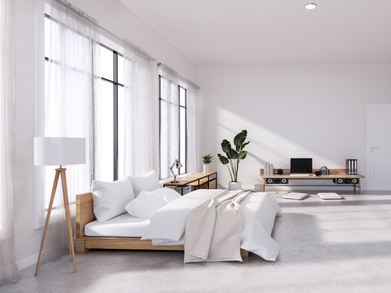 Um quarto minimalista e iluminado com janelas grandes, cortinas transparentes, uma cama baixa de madeira com roupa de cama branca, um abajur, uma planta em vaso e um console de TV com alto-falantes contra uma parede de tijolos brancos.