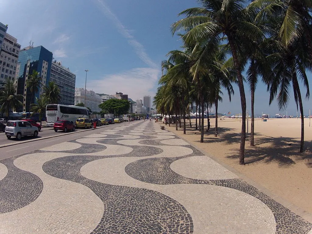 Uma ampla calçada com um padrão de ondas pretas e brancas corre ao longo de uma praia de areia ladeada por palmeiras, capturando o espírito vibrante de Morar no Rio de Janeiro, com carros, prédios e pessoas sob o céu azul.