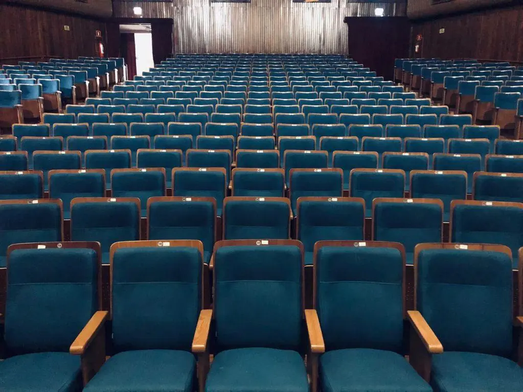 Fileiras de poltronas azuis vazias com braços de madeira dentro do auditório do Teatro Municipal de Ribeirão Preto, todas voltadas para um palco ou tela na frente da sala.