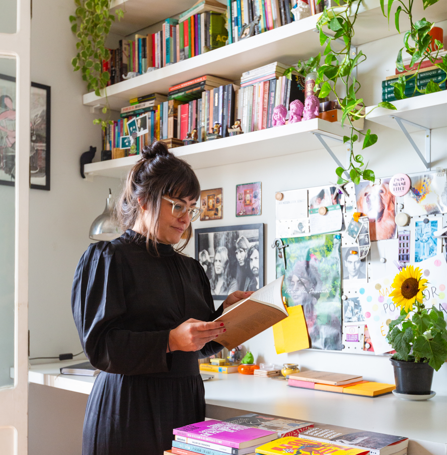 Uma mulher de óculos e vestido preto está no iluminado Apartamento Higienópolis, lendo um livro. Atrás dela, prateleiras repletas de livros e plantas, e uma escrivaninha com cadernos coloridos, girassóis e fotos pregadas na parede.