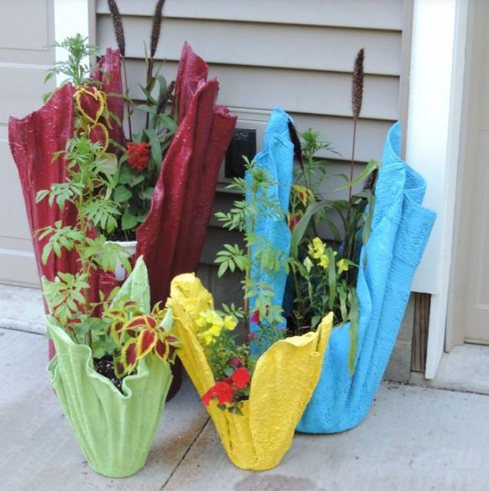 Quatro vasos de cimento coloridos, em formato de tecido, nas cores vermelho, verde, azul e amarelo, estão dispostos em um pátio, encostados na fachada da casa. Cada vaso contém uma variedade de plantas e flores de um verde vibrante.