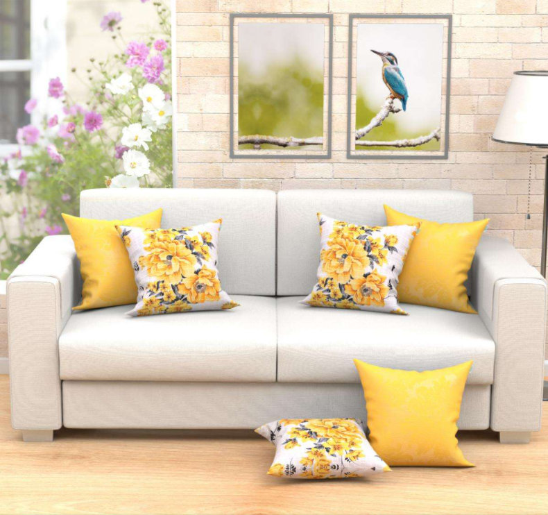 Um sofá cinza-claro com almofadas para sofá em amarelo e estampas florais em uma sala de estar iluminada. Duas almofadas florais e quatro amarelas estão dispostas sobre o sofá e o chão. Arte floral na parede e uma pintura de pássaro estão penduradas acima do sofá.