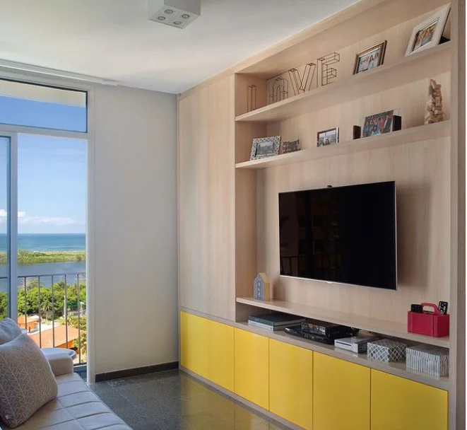 Sala de estar moderna com TV na parede sobre uma estante de madeira, armários inferiores amarelos, itens decorativos e fotos emolduradas. Ideal para decoração de sala pequena, com uma grande janela emoldurando o céu azul, a água e a vegetação lá fora.
