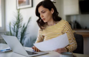 Imagem que ilustra matéria sobre carnê-leão mostra uma mulher sentada em uma cadeira em frente ao computador com um papel em mãos e com uma feição de preocupada