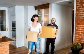 Imagem de um homem e uma mulher entrando em um imóvel, segurando caixas de mudança, para ilustrar matéria sobre casa ou apartamento