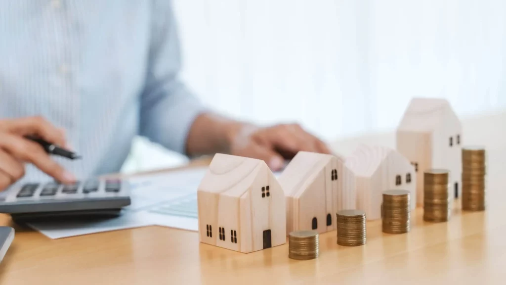 Imagem de um empresário mexendo em um laptop enquanto aponta com uma caneta para moedas e um modelo de casa representando o investimento imobiliário para ilustrar matéria sobre como a CCI funciona no mercado imobiliário