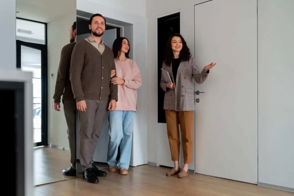 Imagem de uma mulher mostrando um imóvel a um casal sorridente, apontando para um cômodo enquanto está em um corredor moderno e bem iluminado.