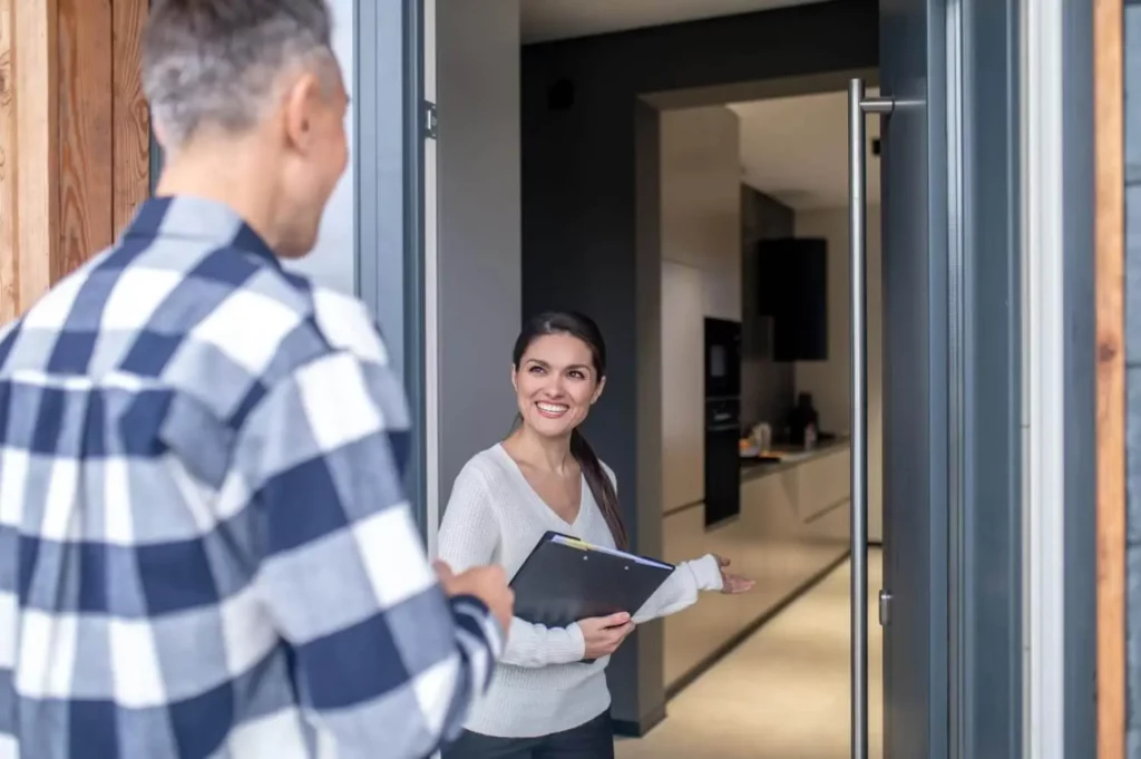 Imagem de uma mulher segurando uma pasta está em pé na entrada aberta, sorrindo e gesticulando para dentro, enquanto um homem de camisa xadrez está do lado de fora, de frente para ela. A imagem mostra uma cozinha moderna ao fundo.