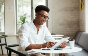 Imagem de um homem de óculos e camisa branca sorrindo enquanto usa um tablet em uma pequena mesa em um café moderno e bem iluminado para ilustrar matéria sobre o que é ITBI. Um smartphone, um notebook e um copo de café descartável estão sobre a mesa.