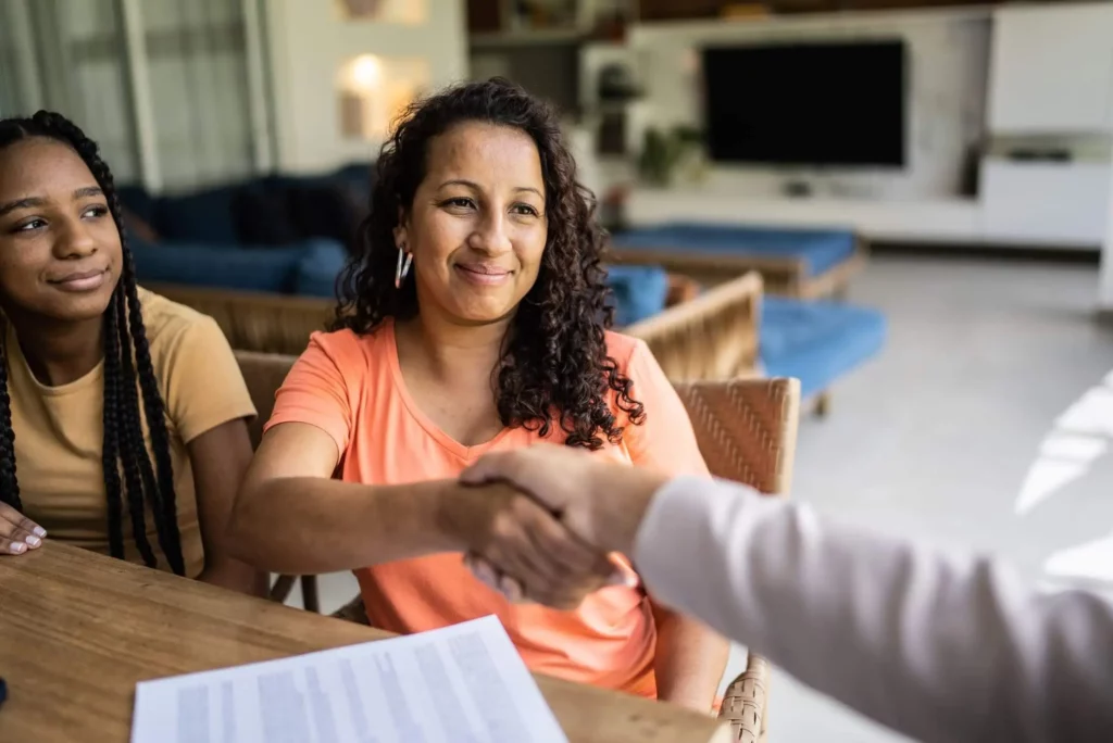 Imagem de duas mulheres sentadas nas cadeiras dispostas perto de uma mesa de madeira enquanto uma delas aperta a mão de um consultor imobiliário para ilustrar matéria que responda a seguinte pergunta: o que devo saber antes de comprar um imóvel?