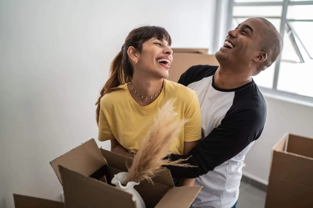 Imagem de um casal composto por um homem e uma mulher sorrindo enquanto abrem uma caixa de papelão para ilustrar matéria que responda a seguinte pergunta: o que devo saber antes de comprar uma casa?