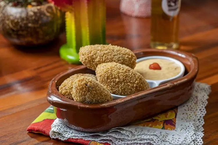 Quatro croquetes empanados e fritos são servidos em um prato de cerâmica marrom com molho cremoso e uma porção de molho picante, que lembra os sabores que você encontraria no Centro de Tradições Nordestinas, tudo sobre um guardanapo de renda sobre uma mesa de madeira.