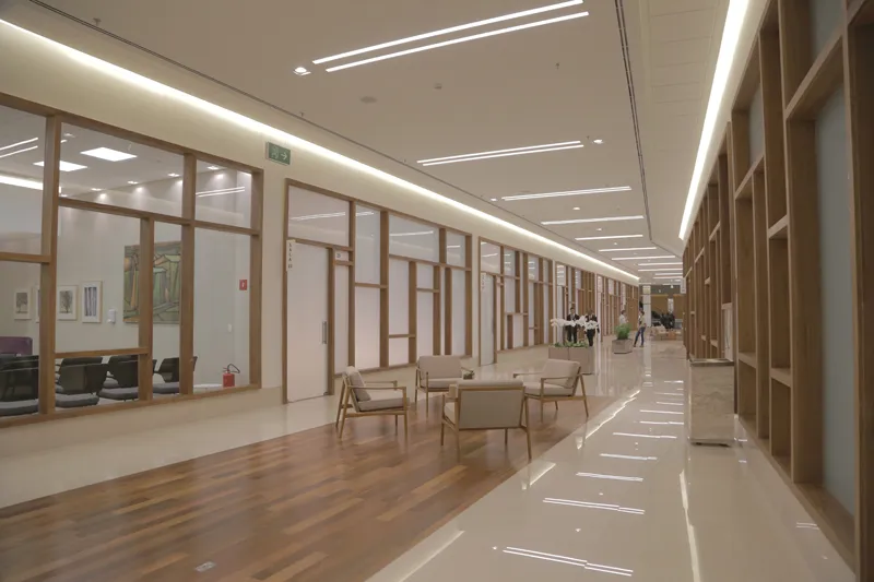Um corredor moderno e espaçoso, inspirado no Ribeirão Shopping, com grandes janelas de vidro, detalhes em madeira e uma área de estar com cadeiras bege e uma mesa. O corredor é bem iluminado e tem piso polido.