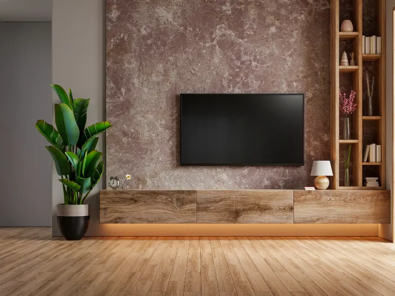 Uma sala de estar moderna conta com uma TV de tela plana montada na parede, um console de madeira com decoração, uma planta alta em um vaso preto, prateleiras embutidas e um elegante papel de parede para sala na parede marrom texturizada sobre o piso de madeira.