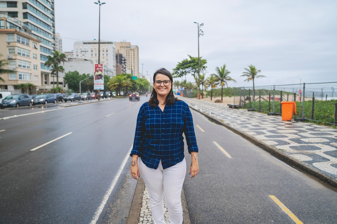 Uma mulher usando óculos, camisa xadrez azul e calça branca está sorrindo em uma rua molhada na praia de Ipanema, cercada por palmeiras, carros, prédios e céu nublado — mostrando o charme de morar em Ipanema.