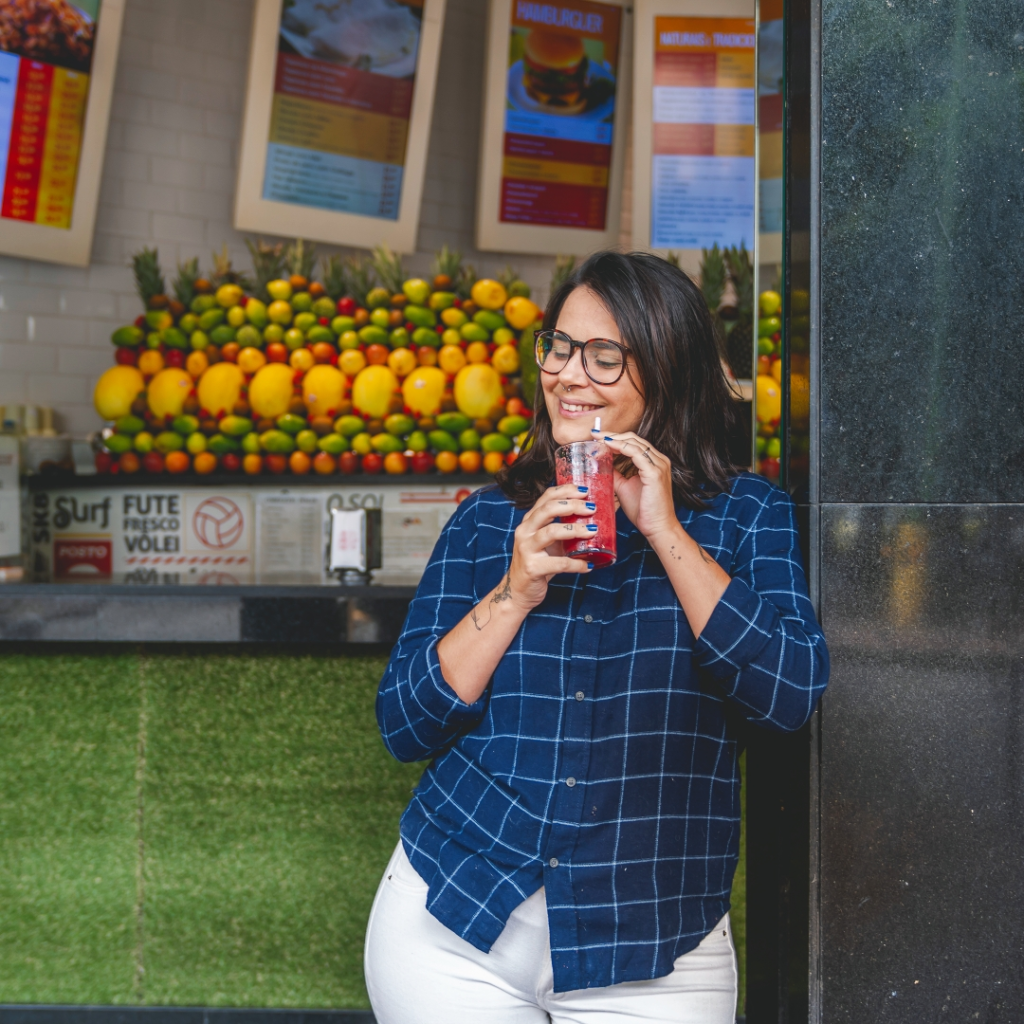 Uma mulher de óculos, camisa xadrez azul e calça branca sorri enquanto bebe um suco vermelho do lado de fora de um bar de sucos de frutas colorido, curtindo o clima de morar em Ipanema.