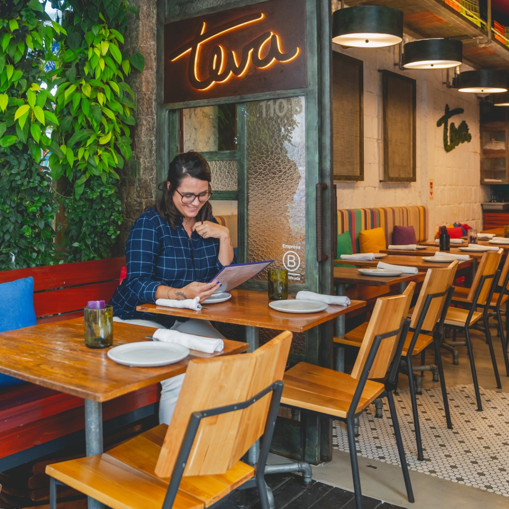 Uma mulher sentada sozinha a uma mesa de madeira em um restaurante colorido e aconchegante em Ipanema, sorrindo enquanto olha o cardápio. A iluminação aconchegante, as plantas exuberantes e uma placa de neon da Tava na porta de vidro capturam o charme do "Morar em Ipanema".
