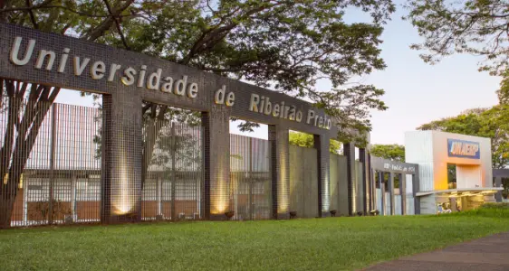 Placa de entrada da Universidade de Ribeirão Preto (UNAERP), uma das principais faculdades de Ribeirão Preto, com letras grandes em um muro de pedra, árvores atrás e um prédio branco da UNAERP visível à direita.