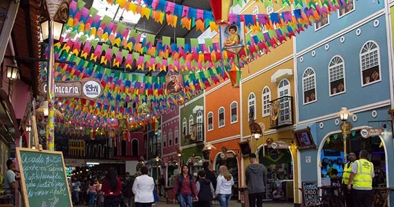 Uma animada cena de rua no Centro de Tradições Nordestinas mostra pessoas caminhando sob bandeiras de papel coloridas, com edifícios vibrantes e multicoloridos alinhados em ambos os lados e decorações festivas criando uma atmosfera alegre.