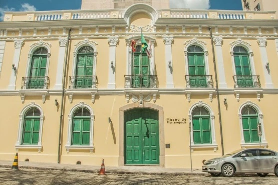 O Museu de Florianópolis é um prédio histórico amarelo e branco com portas e janelas verdes, que ostenta orgulhosamente bandeiras acima de sua entrada. Um carro prateado e cones de trânsito podem ser vistos em frente ao museu.