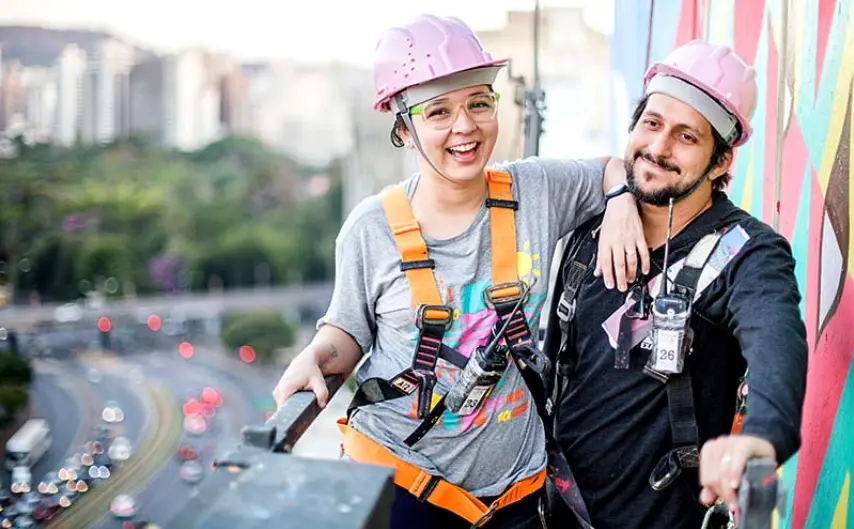 Duas pessoas usando cintos de segurança e capacetes rosa sorriem em uma saliência alta ao lado do vibrante grafite em BH, com uma paisagem urbana e uma estrada movimentada ao fundo.