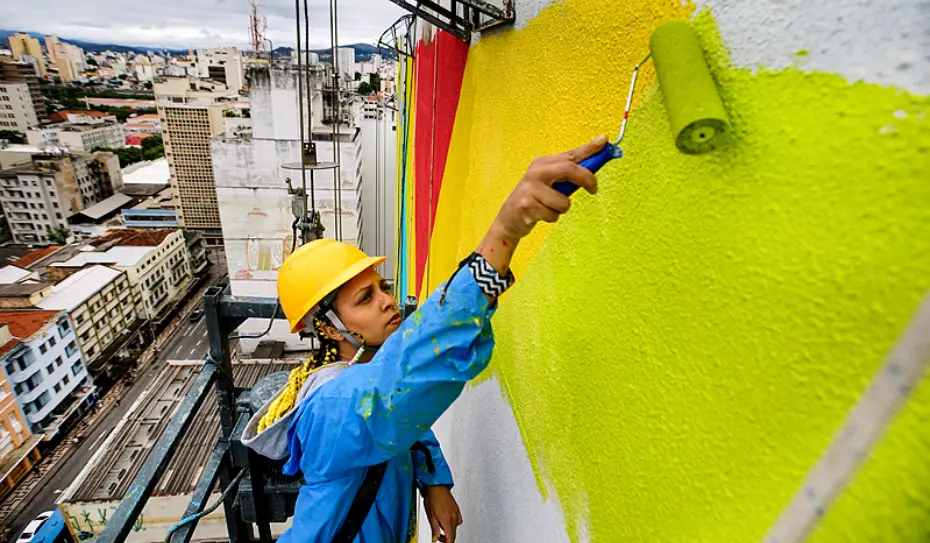 Uma pessoa usando um capacete amarelo e uma jaqueta azul usa um rolo de pintura para adicionar amarelo brilhante sobre grafite em BH, em pé em um andaime bem acima da paisagem urbana.