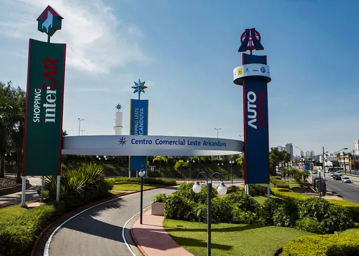 Entrada do Centro Comercial Leste Aricanduva, em São Paulo, Brasil, com placas destacadas do Shopping Aricanduva, área verde exuberante e uma rotatória na estrada principal sob um céu limpo.