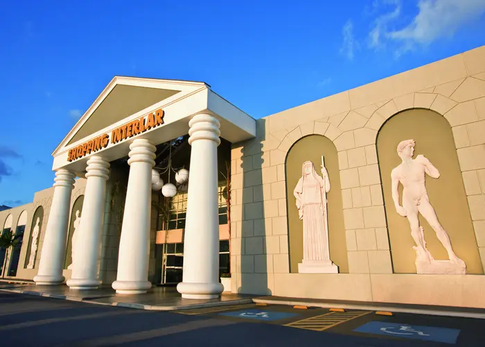 Shopping center com altas colunas brancas, uma grande entrada de vidro e estátuas clássicas — incluindo uma réplica do David de Michelangelo — dispostas em nichos ao longo da fachada bege sob um céu azul claro, que lembra o estilo grandioso do shopping Aricanduva.